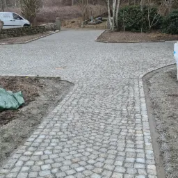 Ukończona podjazd z kostki granitowej w kształcie wachlarza, widoczne materiały budowlane, w tle budynek i zaparkowany samochód dostawczy.