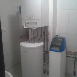 Biała pompa ciepła z zasobnikiem i zmiękczaczem wody w kafelkowanej kotłowni. Widoczne rury miedziane i elementy instalacji. Oświetlenie górne.