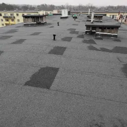 Szary, płaski dach budynku po naprawie z widocznymi łatami i kominami. W tle budynki mieszkalne i pochmurne niebo.