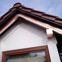 Dach-Pol - Detal wykończenia dachu z miedzianymi obróbkami blacharskimi, widoczne dachówki i fragment elewacji z oknem. Ujęcie z dołu, na tle błękitnego nieba.