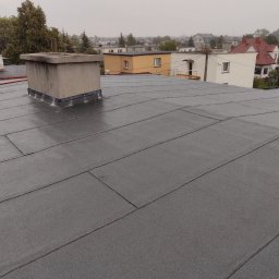 Dach-Pol - Szary, płaski dach pokryty papą bitumiczną z kominem w tle zabudowy miejskiej w pochmurny dzień. Widok z góry na nową izolację dachu.
