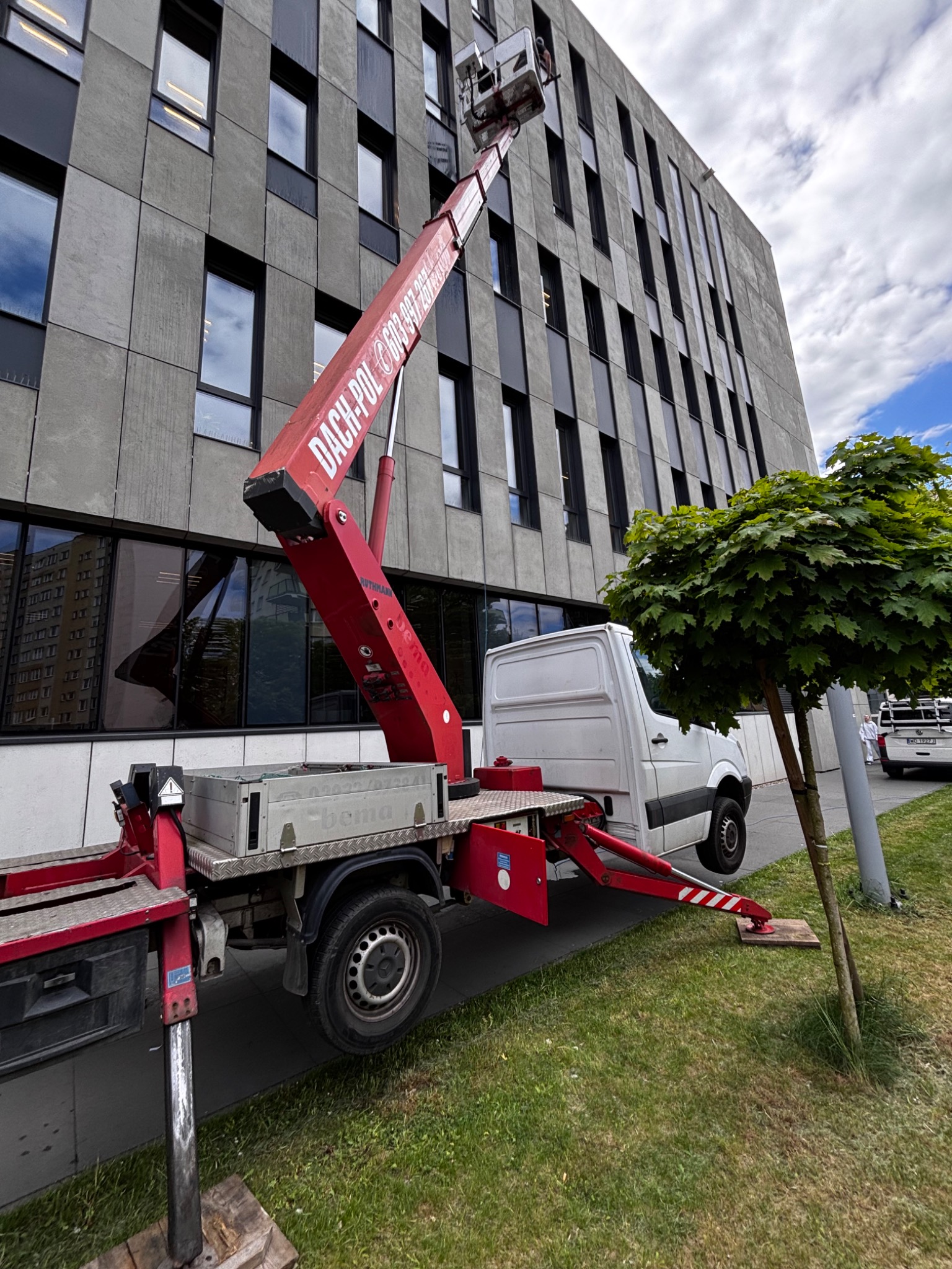 Czerwony podnośnik koszowy Dach-Pol na białym samochodzie dostawczym na tle nowoczesnego biurowca z szarymi panelami i licznymi oknami.