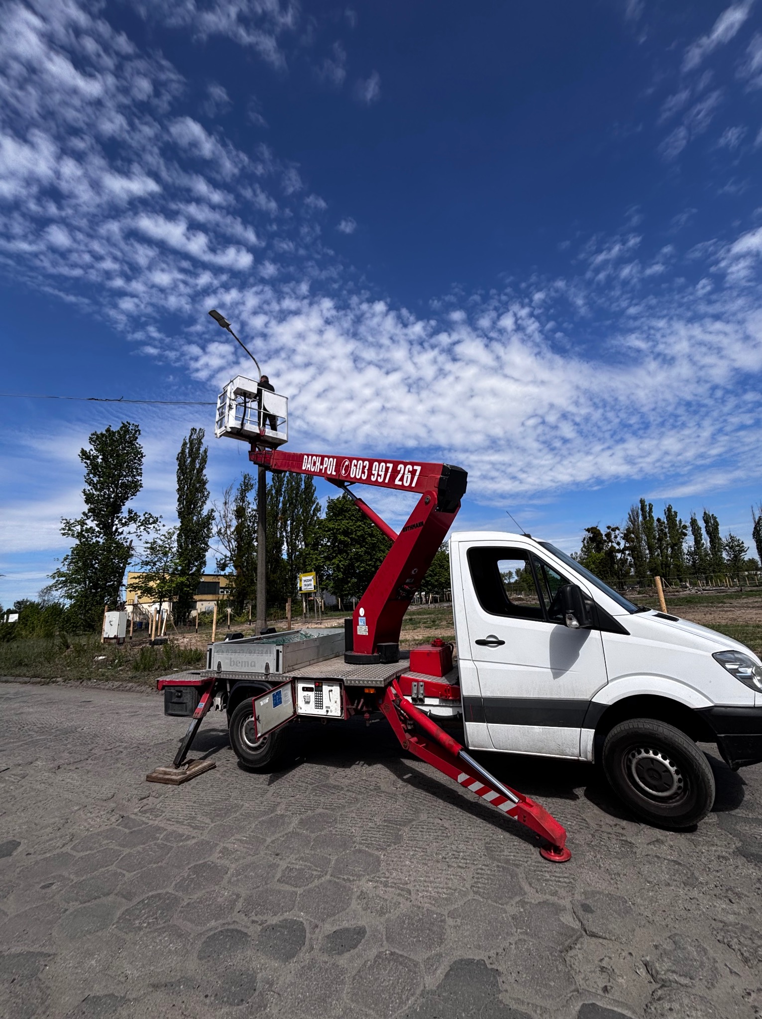 Biały samochód z podnośnikiem koszowym DACH-POL, numer telefonu na czerwonym ramieniu. Operator w koszu, błękitne niebo z chmurami w tle. Usługi dźwigowe.
