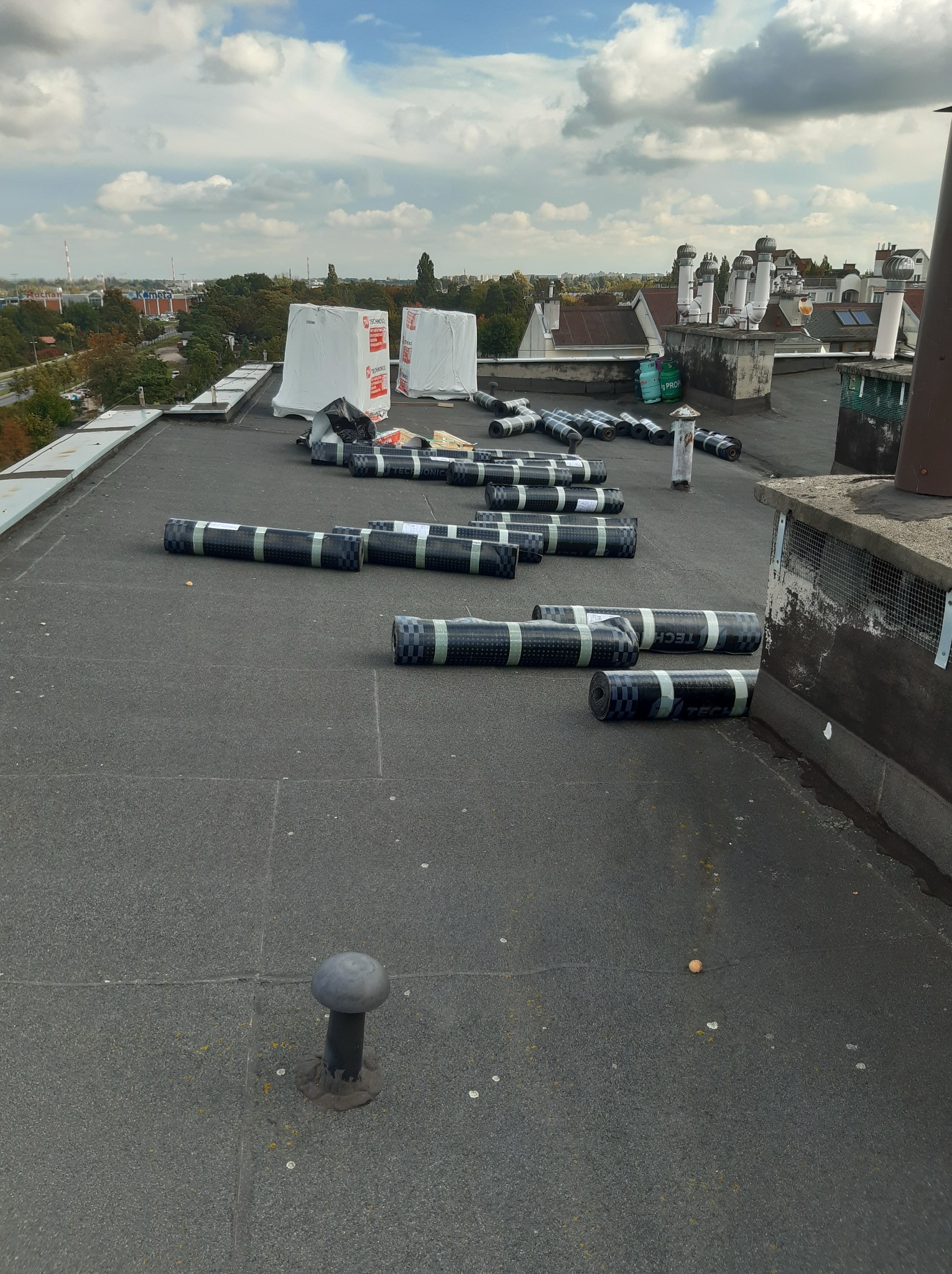 Rozwinięte rolki papy na płaskim dachu przed montażem, widok z góry, w tle zabudowa miejska i pochmurne niebo. Część rolek zabezpieczona folią.