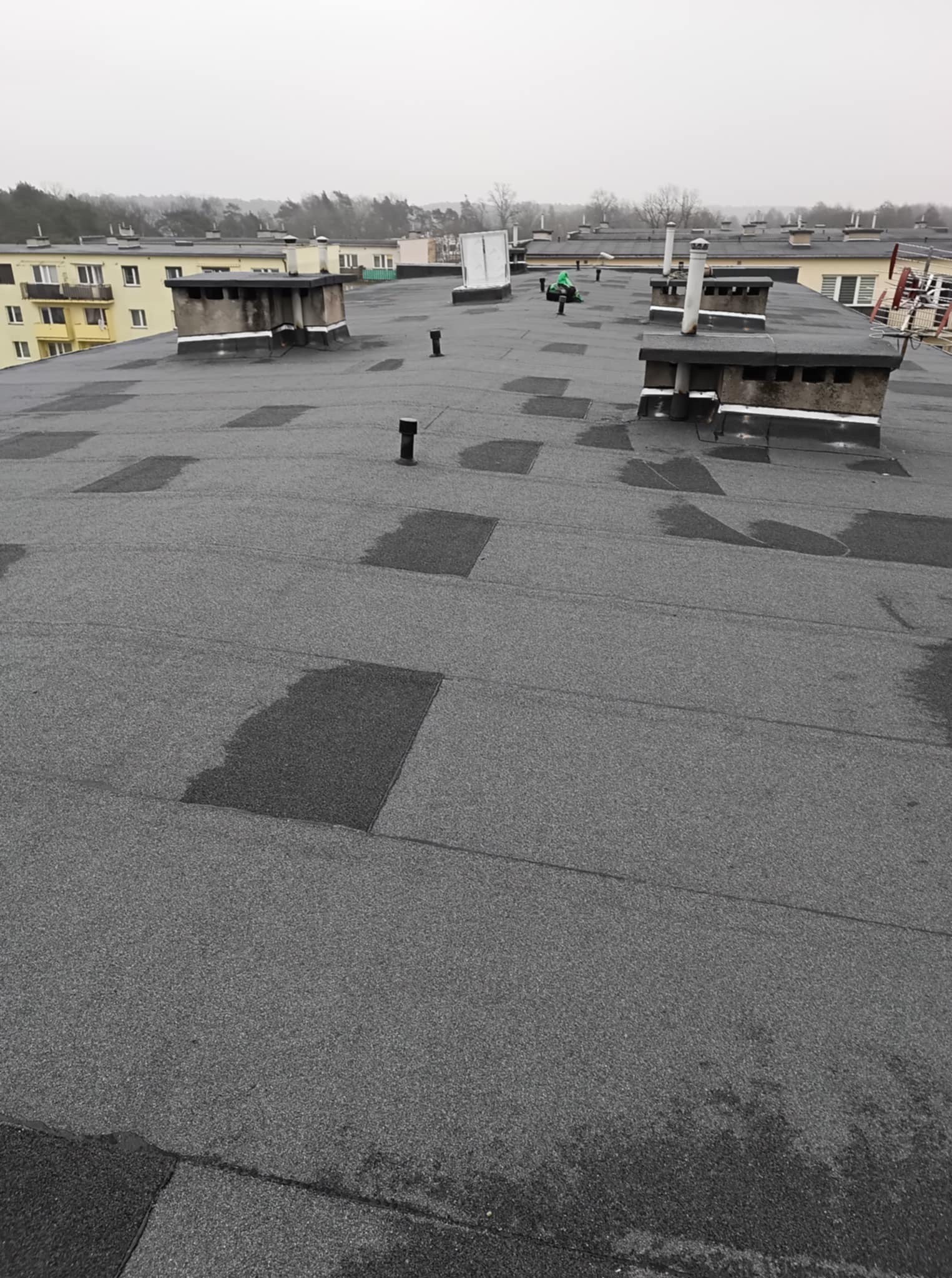 Szary, płaski dach budynku po naprawie z widocznymi łatami i kominami. W tle budynki mieszkalne i pochmurne niebo.