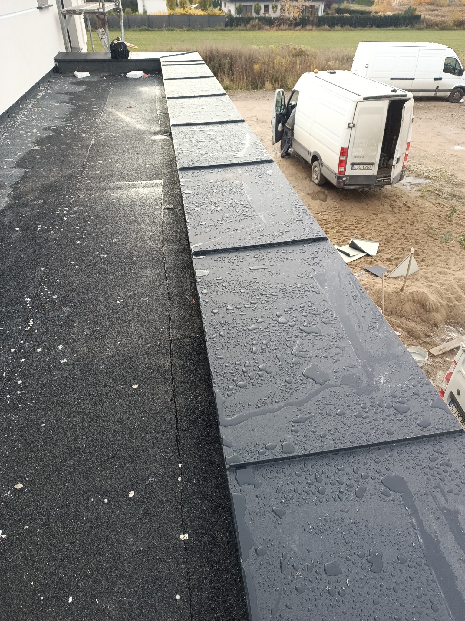 Wodoodporne, grafitowe panele dachowe z widocznymi kroplami deszczu, ułożone na czarnym, bitumicznym podłożu. W tle dwa białe busy i budynek w budowie.