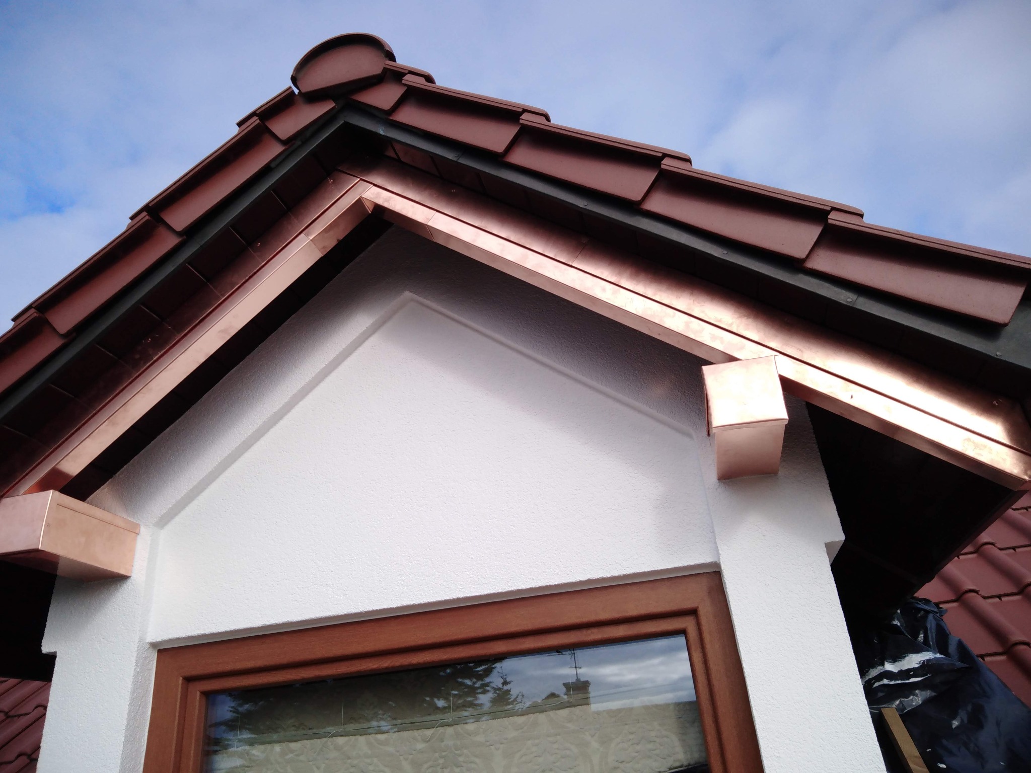 Detal wykończenia dachu z miedzianymi obróbkami blacharskimi, widoczne dachówki i fragment elewacji z oknem. Ujęcie z dołu, na tle błękitnego nieba.