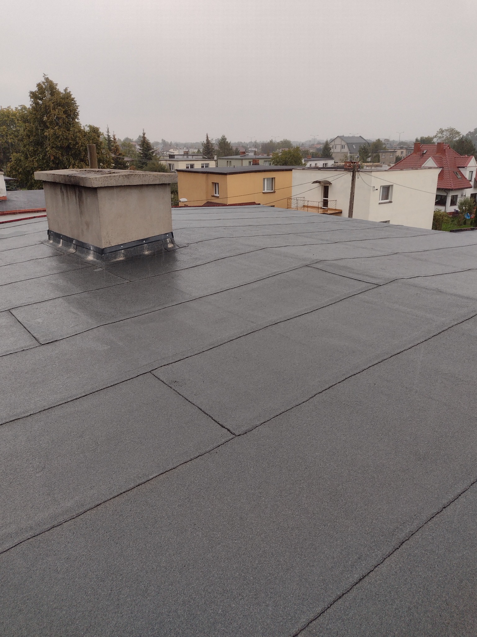 Szary, płaski dach pokryty papą bitumiczną z kominem w tle zabudowy miejskiej w pochmurny dzień. Widok z góry na nową izolację dachu.