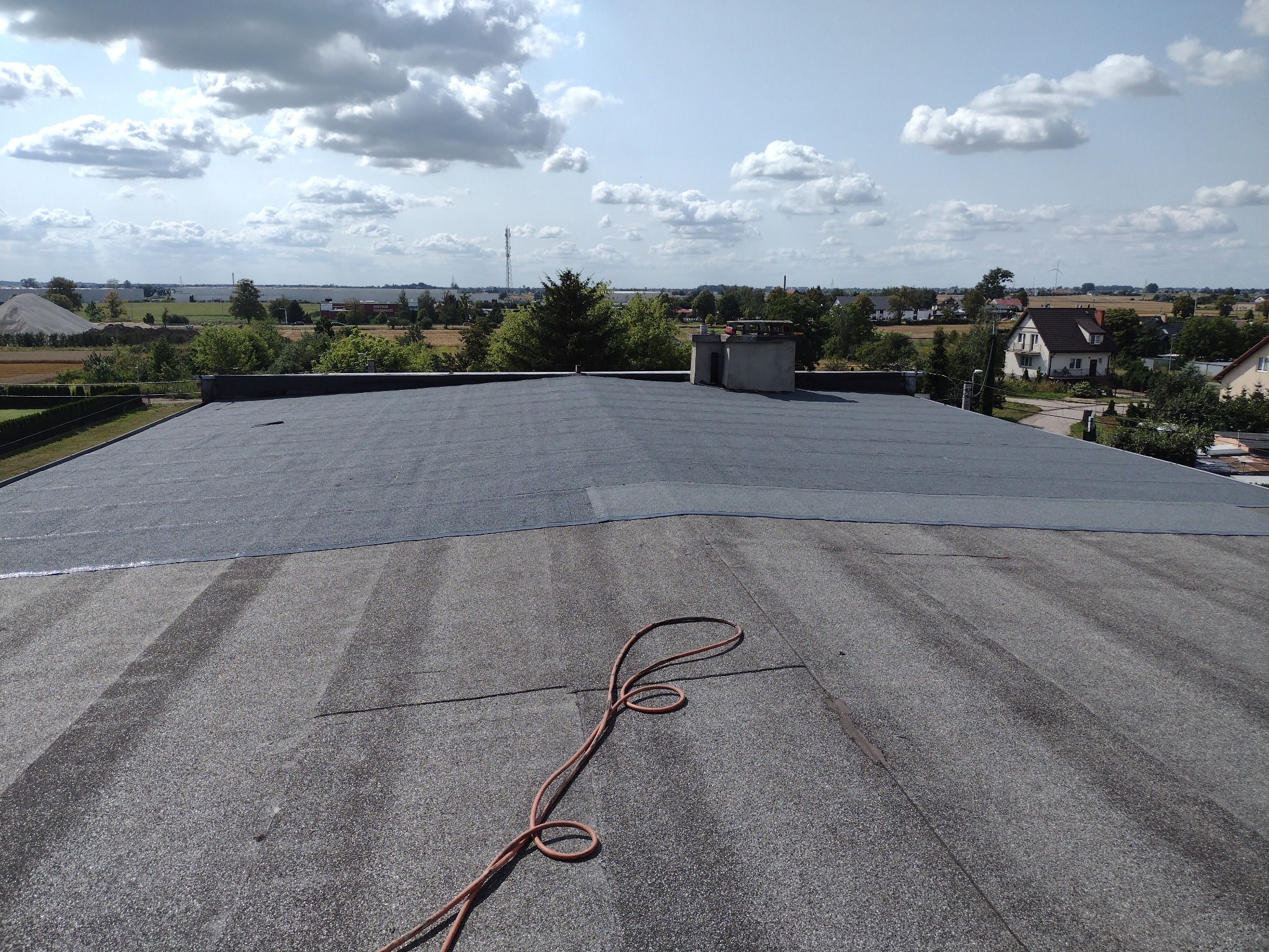 Wykończony, płaski dach pokryty papą termozgrzewalną, z widocznym kominem i przewodem. W tle zabudowa wiejska i błękitne niebo z chmurami.