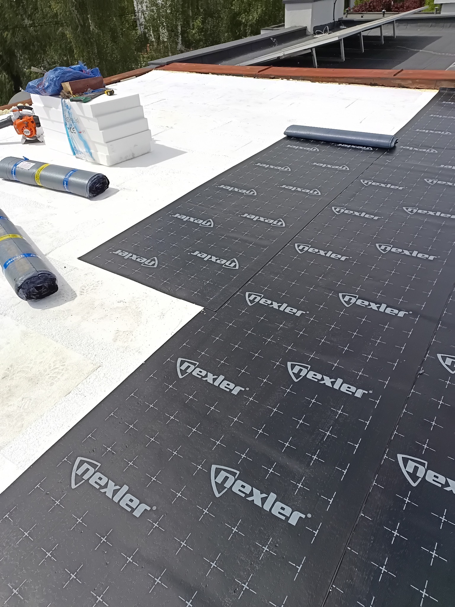 Białe płyty izolacyjne i czarna membrana dachowa Nexler na płaskim dachu. Częściowo rozłożona rolka membrany, narzędzia i materiały budowlane w tle.
