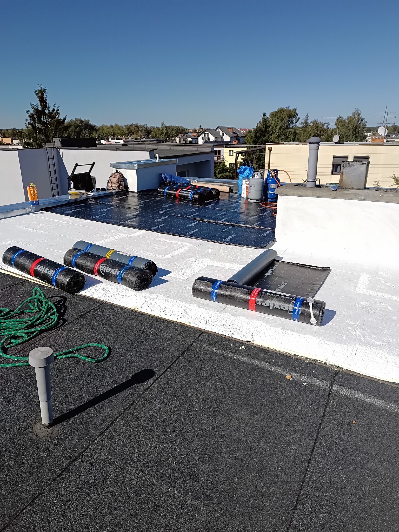 Wykończony dach płaski z czarną papą, częściowo pokryty nową izolacją. Widoczne rolki papy, butle gazowe i narzędzia dekarskie. Słoneczny dzień.