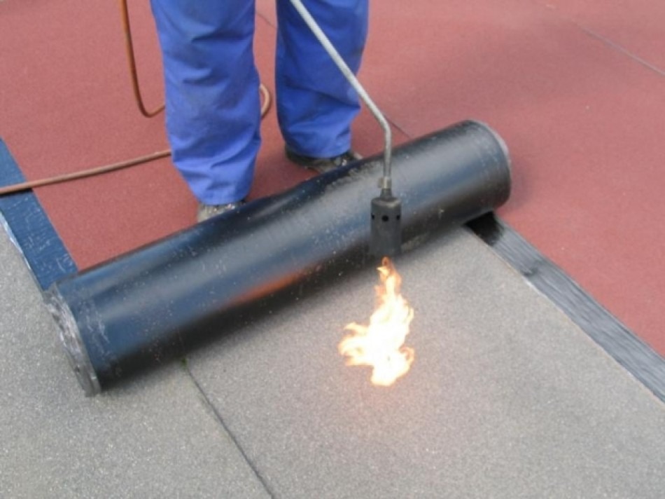 Pracownik w niebieskich spodniach zgrzewa papę termozgrzewalną na dachu za pomocą palnika; widoczny płomień palnika i częściowo rozłożona rolka papy.