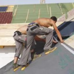 Dwóch mężczyzn bez koszulek, w szarych spodniach roboczych i butach, instaluje czarną membranę dachową z zielonymi napisami na dachu w trakcie budowy. Widoczne drewniane krokwie i fragmenty innych...