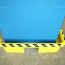 Bariera ochronna w kolorze żółtym z czarnymi pasami, zabezpieczająca niebieską ścianę. Bariera wykonana z tworzywa sztucznego, zamontowana przy podłodze.