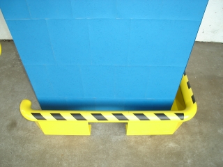 Bariera ochronna w kolorze żółtym z czarnymi pasami, zabezpieczająca niebieską ścianę. Bariera wykonana z tworzywa sztucznego, zamontowana przy podłodze.