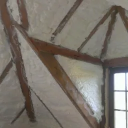 Poddasze z drewnianą konstrukcją pokryte białą pianą izolacyjną, widoczne okno. Izolacja natryskowa poddasza.