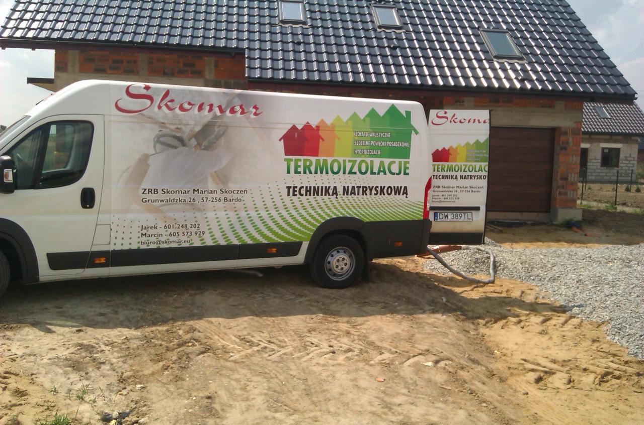 Pojazd firmy Skomar z reklamą termoizolacji natryskowej na tle domu z ciemną dachówką i oknami dachowymi. Widoczne logo, dane kontaktowe i hasła reklamowe.