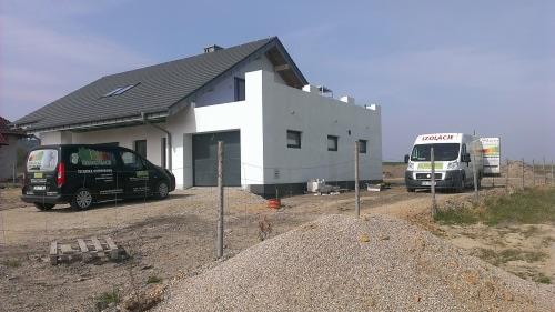 Dom w trakcie prac elewacyjnych, widoczne samochody dostawcze z logo firmy, częściowo ocieplona ściana szczytowa, przygotowany teren.