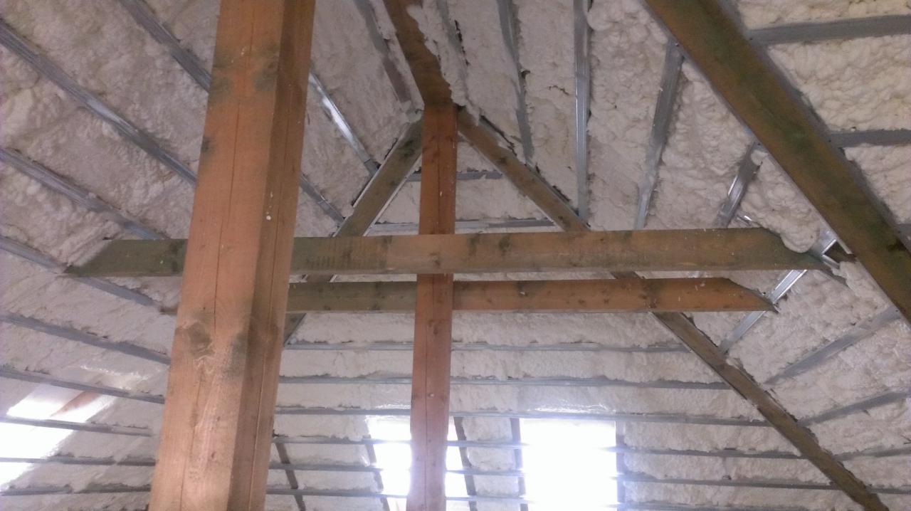 Poddasze z drewnianą konstrukcją i szarą, metalową kratownicą wypełnioną białą pianką izolacyjną. Widoczne belki drewniane i fragment okna dachowego.