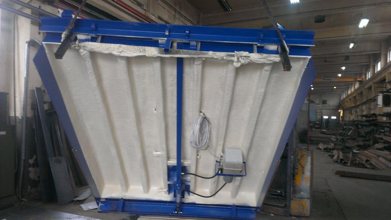 Ocieplony od wewnątrz metalowy element konstrukcyjny w hali produkcyjnej. Widoczne niebieskie wzmocnienia i biała pianka izolacyjna. Element zawieszony na hakach.