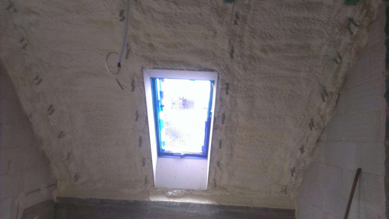 Poddasze z oknem dachowym po natrysku pianki izolacyjnej, widoczne mocowania i fragment instalacji elektrycznej. Ściany wykończone pianą, okno zabezpieczone folią.