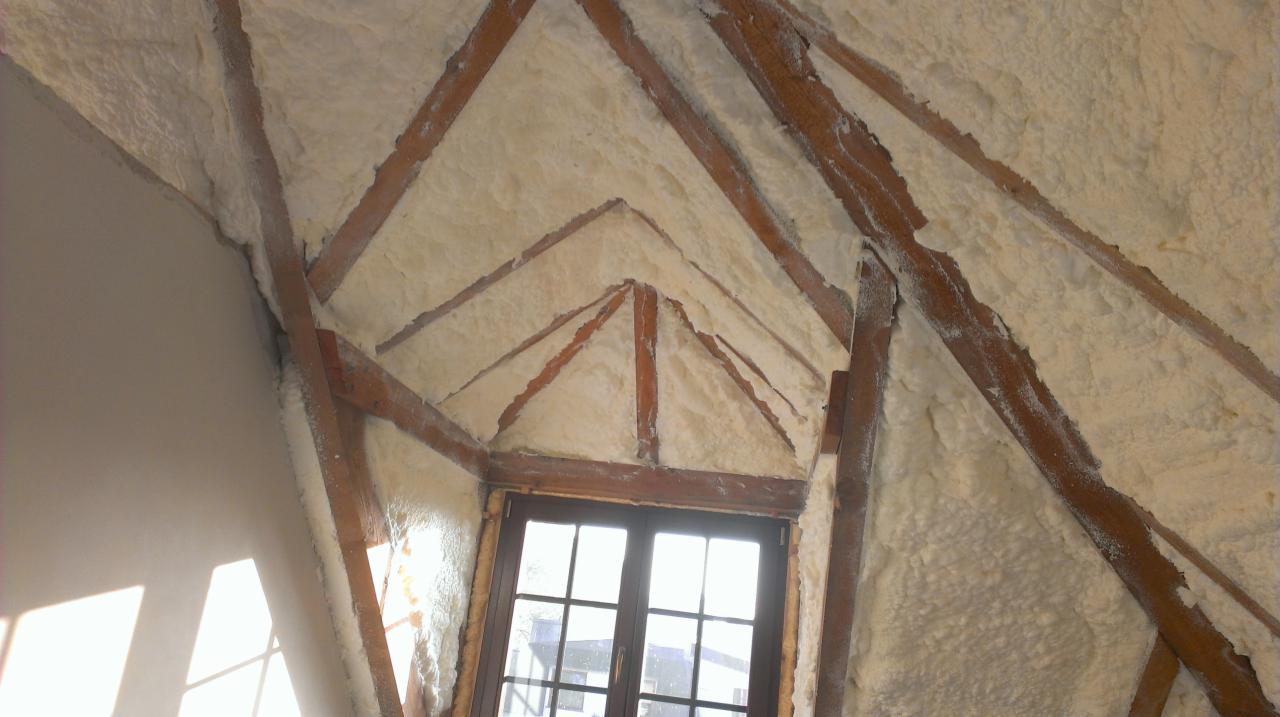 Poddasze z drewnianą konstrukcją pokryte pianą izolacyjną, widoczne okno. Ocieplenie poddasza pianką poliuretanową, detale drewnianych belek. Jasne, naturalne oświetlenie.