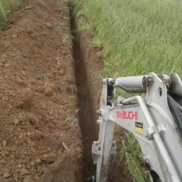 Wykopany wąski rów w ziemi, widoczna część minikoparki Takeuchi, w tle trawa i dom.