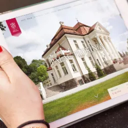 Kobieta trzyma tablet prezentujący stronę internetową Pałacu Tłokinia, z datami rezerwacji widocznymi na ekranie.