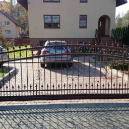 Automatyczna brama wjazdowa z metaloplastyki w kolorze ciemnego brązu, z dekoracyjnymi elementami, zamykająca wjazd na posesję z kostki brukowej, w tle dom jednorodzinny i zaparkowany samochód.