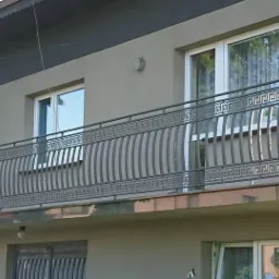 Szara, metalowa balustrada balkonowa z motywem greckim na tle jasnego budynku mieszkalnego, widok z dołu.
