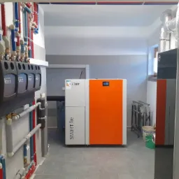 Pomieszczenie kotłowni z zainstalowanym piecem na paliwo stałe Smart Fire Lazar z pomarańczowym frontem, zasobnikiem Altech i rozbudowaną instalacją hydrauliczną z rurami w kolorach czerwonym...