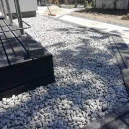 Wykończona ścieżka z jasnych kamieni prowadząca od drewnianego tarasu do chodnika z kostki brukowej, widoczna sterta piasku i narzędzia w tle.