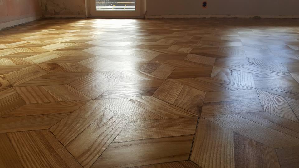 Wypolerowany parkiet ułożony we wzór jodełki francuskiej, widoczne refleksy światła z okna, ściany w trakcie remontu.