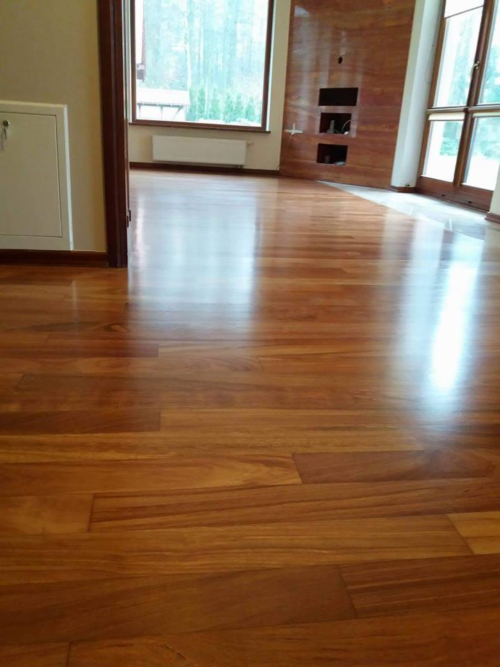 Błyszcząca, świeżo wykończona drewniana podłoga w przestronnym salonie z dużymi oknami i drewnianą ścianą akcentującą.