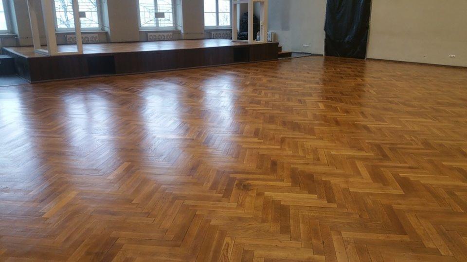 Sala z parkietem ułożonym w jodełkę po renowacji, widoczny połysk lakieru, w tle scena i okna.