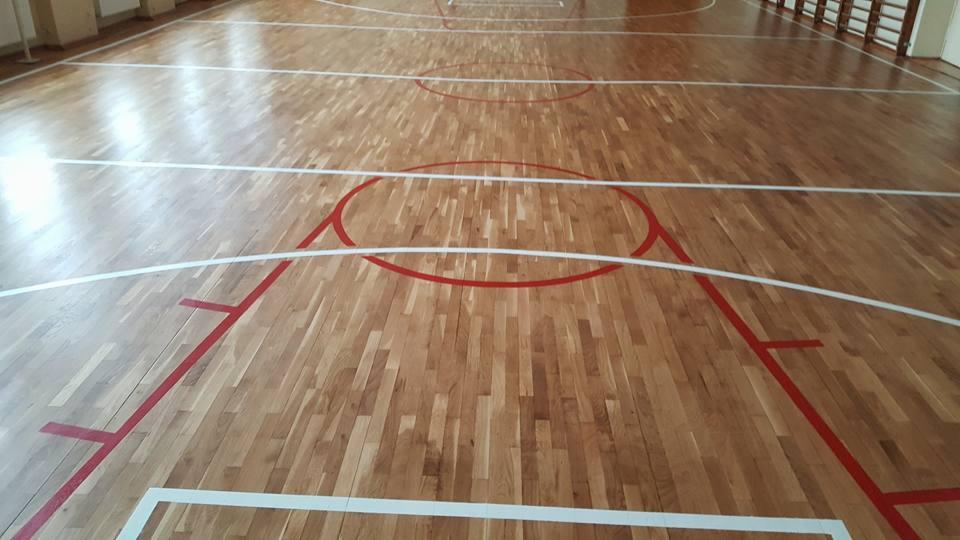 Parkiet z drewna liściastego na sali gimnastycznej z czerwonymi i białymi liniami boiska do koszykówki, widoczne ślady po zakończeniu prac renowacyjnych.