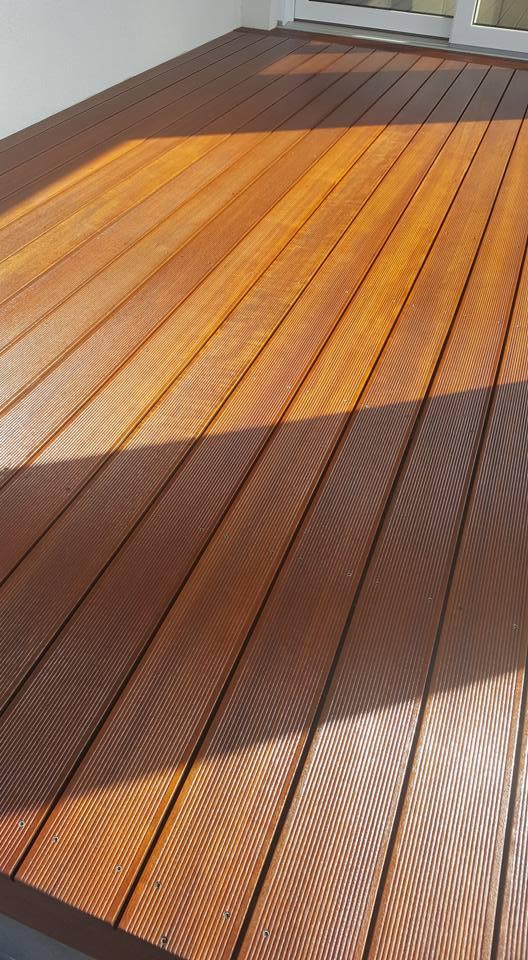 Świeżo polakierowany parkiet z egzotycznego drewna w ciepłym odcieniu brązu, z widocznymi delikatnymi rowkami i odbiciem światła słonecznego.