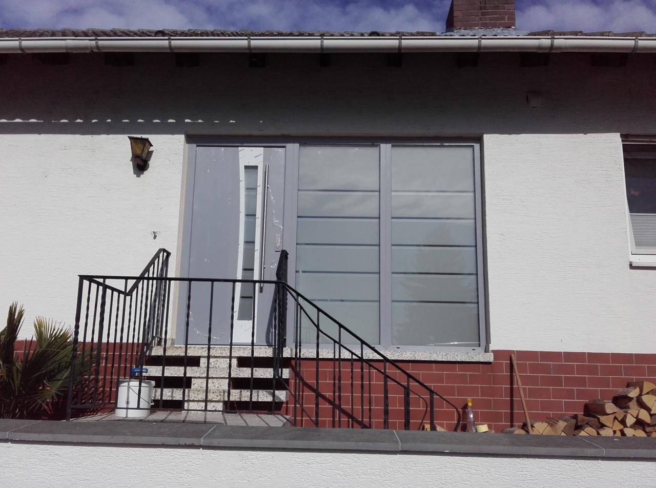 Wejście do domu z szarą stolarką okienną i drzwiową, widoczne schody z czarną balustradą, fragment elewacji z fakturą baranka oraz stos drewna przy ścianie.