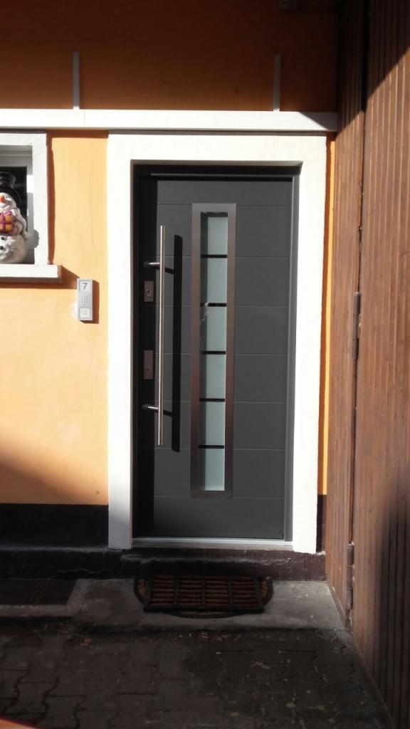 Nowoczesne, grafitowe drzwi wejściowe z przeszkleniami i długim, pionowym uchwytem, zamontowane w pomarańczowej ścianie z białym obramowaniem, widoczny numer domu nad dzwonkiem.