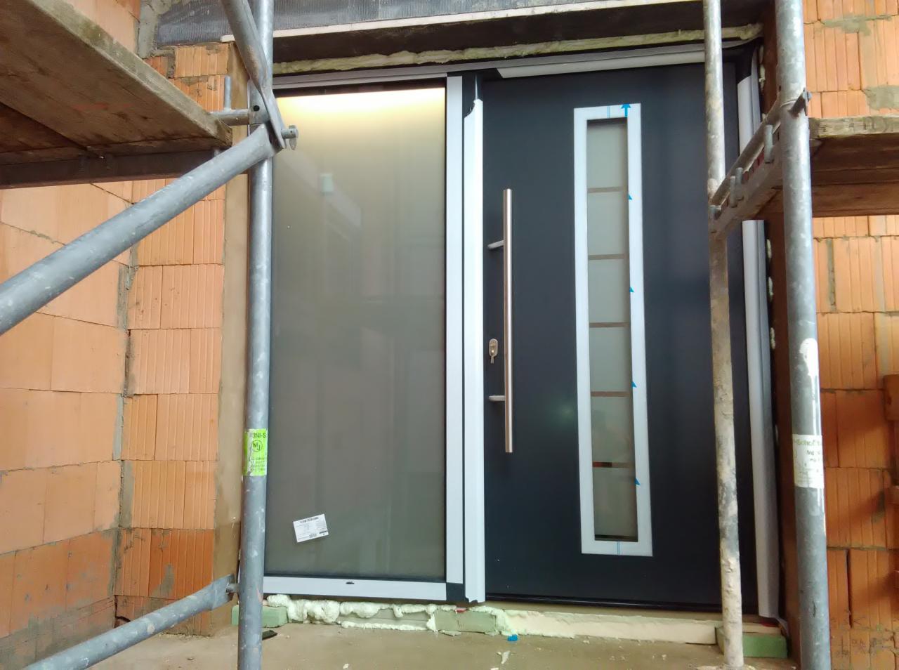 Montaż nowoczesnych drzwi wejściowych z przeszkleniami w surowym stanie, widoczne rusztowania i elementy konstrukcyjne budynku.