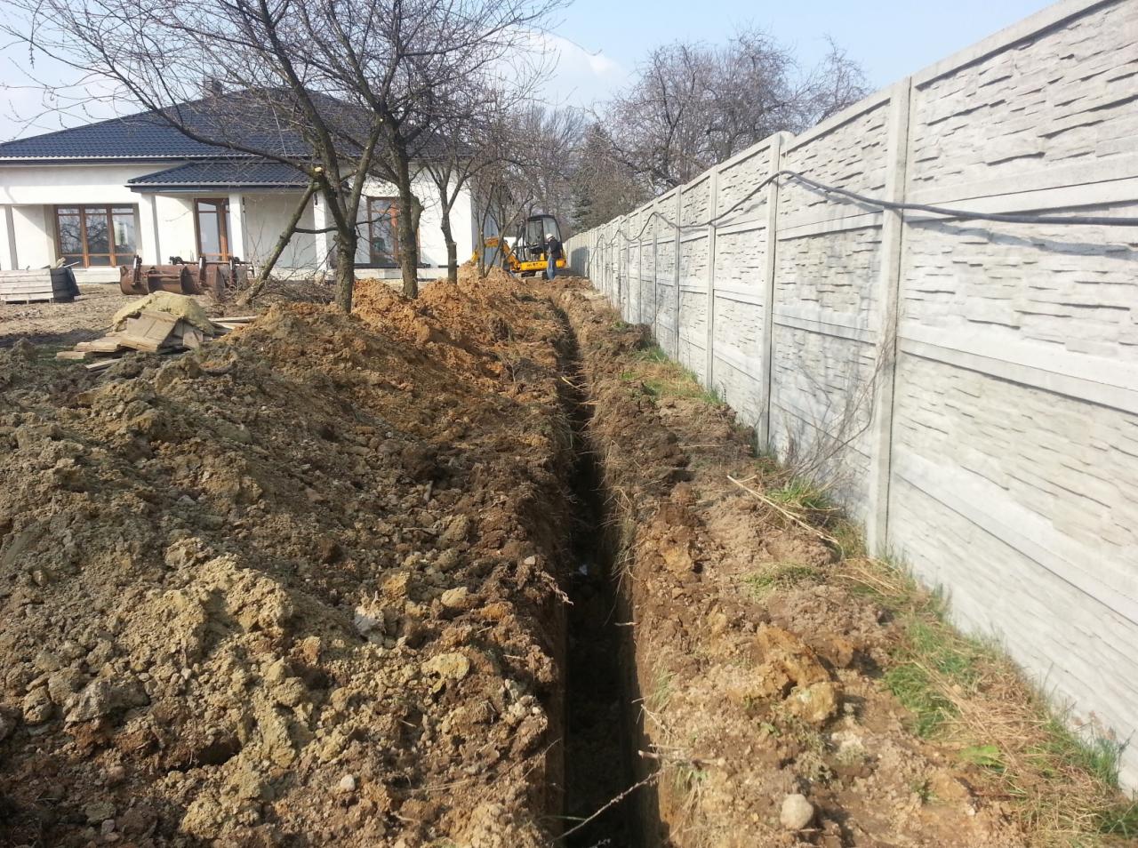 Długi wykop w ziemi wzdłuż betonowego ogrodzenia, w tle dom jednorodzinny i mała koparka.