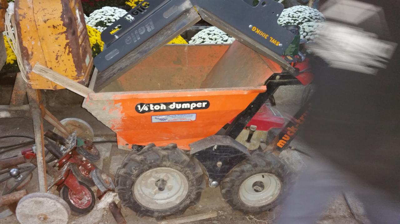 Pomarańczowa wywrotka 1/4 ton dumper marki Muck-Truck, zaparkowana obok żółtego zbiornika i kwiatów doniczkowych.