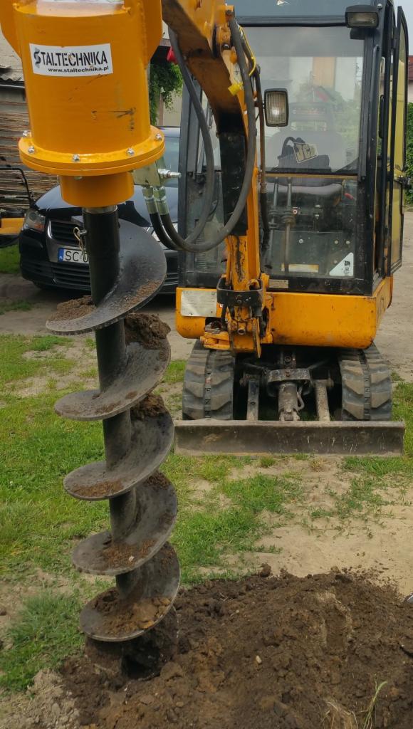 Żółta wiertnica zamontowana na małej koparce, wierci otwór w ziemi, z widocznym logo 'Staltechnika' na górze wiertnicy.