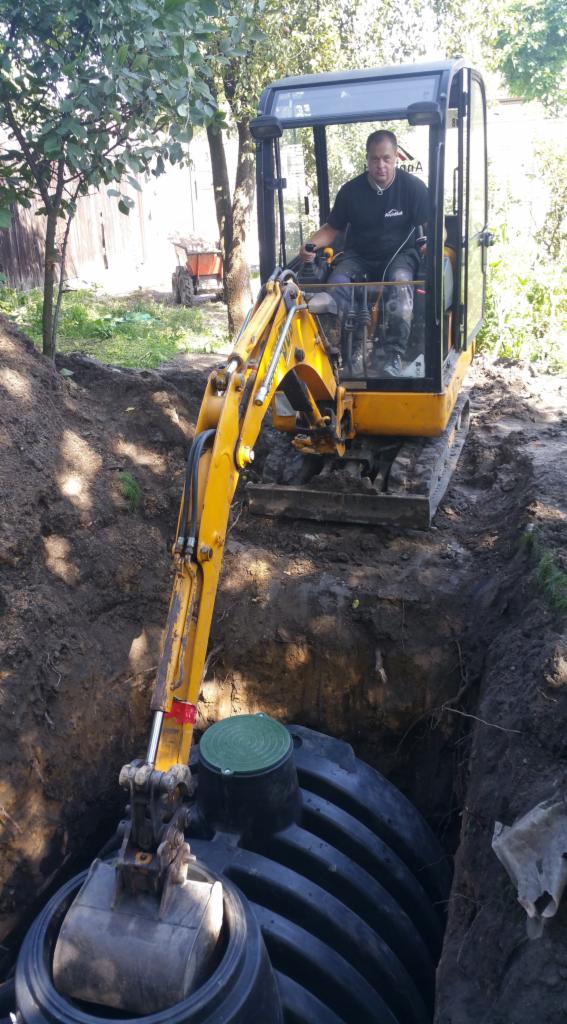 Żółta mini koparka umieszcza plastikowy zbiornik z zieloną pokrywą w wykopie ziemnym; operator w kabinie, w tle drzewa i ogrodzenie.
