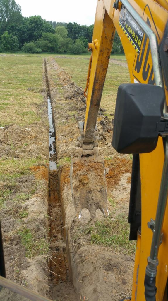 Żółta koparka wykopuje wąski rów w ziemi na łące, z wodą zbierającą się na dnie rowu.