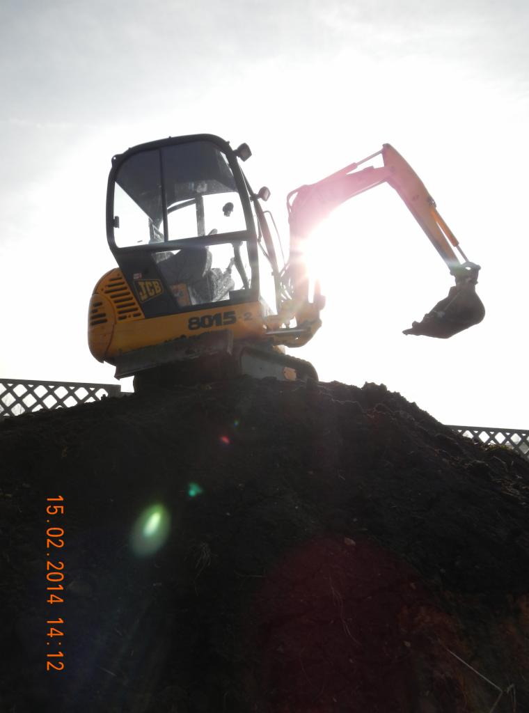 Żółta minikoparka JCB 8015-2 na szczycie pryzmy ziemi pod ostrym słońcem, z widocznym fragmentem ogrodzenia w tle.