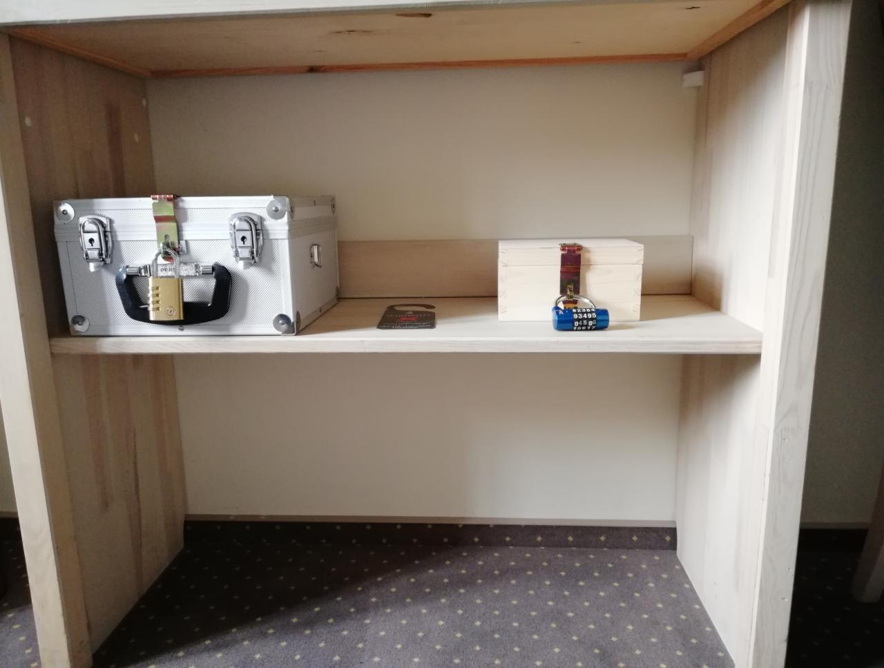 Pokój zagadek: metalowa walizka i drewniane pudełko zabezpieczone kłódkami szyfrowymi na półce w drewnianej szafce.