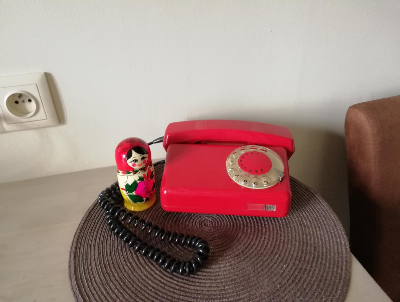 Czerwony telefon stacjonarny z tarczą obrotową i drewniana lalka matrioszka stojące na okrągłej podkładce na stole.