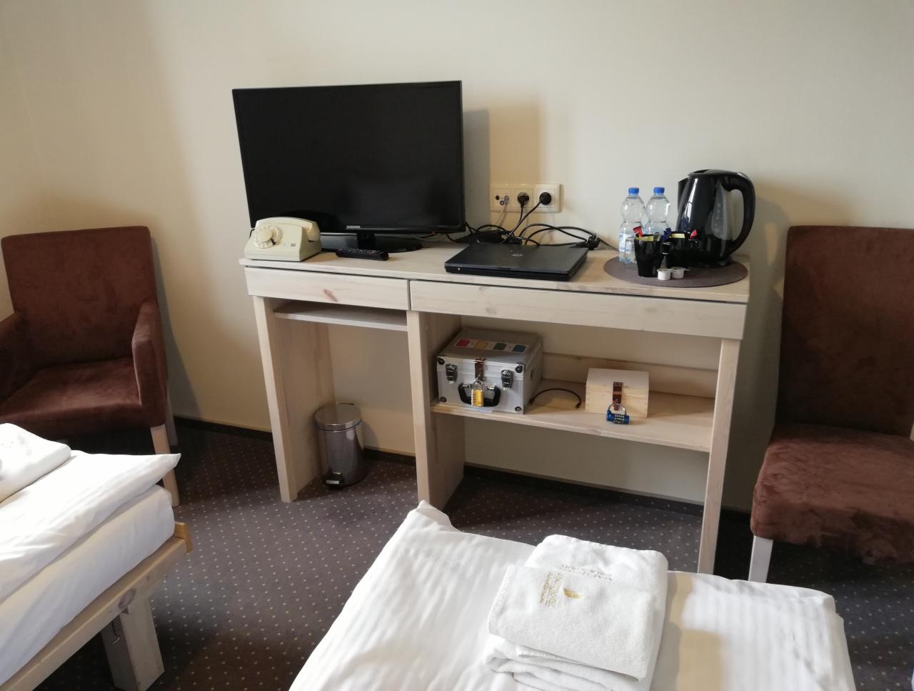 Pokój hotelowy z dwoma łóżkami, biurkiem z telewizorem, czajnikiem, telefonem retro i metalową skrzynką na półce.