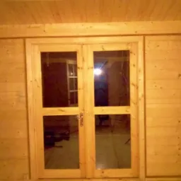 Wnętrze domu wykończone drewnem, z podwójnymi przeszklonymi drzwiami na środku, ozdobnymi kinkietami po bokach i drewnianymi komodami po bokach, oświetlone ciepłym światłem.